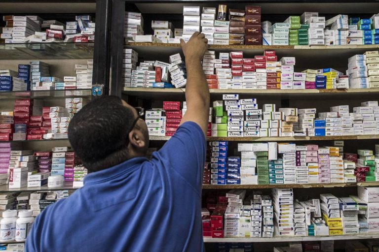 84288-768x512 | MCG24 | موقع إلكتروني مغربي شامل متجدد على مدار الساعة A person in a blue shirt reaches up to grab a box from high pharmacy shelves full of medicine boxes.