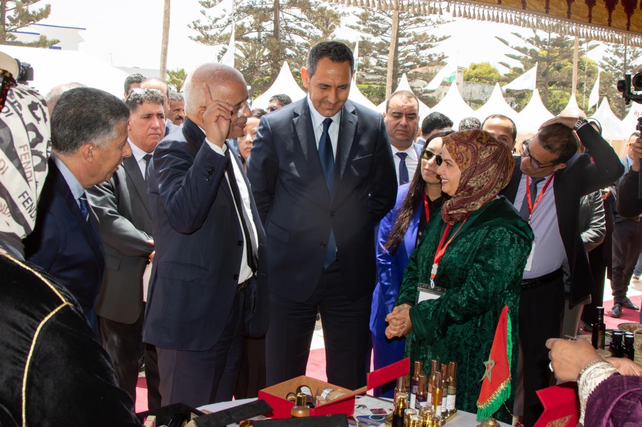 Dignitaries in suits listen as a man gestures, a woman in a green dress and hijab speaks at an outdoor expo booth with perfume bottles on display and a Moroccan flag nearby.