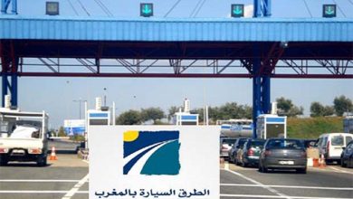 Toll plaza on a highway with several lanes and cars, blue structures, and a sign reading 'Autoroutes du Maroc' in Arabic and French.