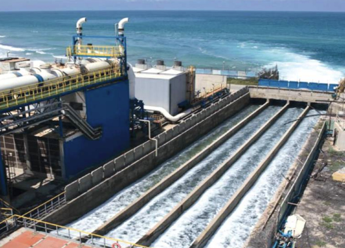 Industrial water treatment plant with open troughs carrying flowing water, adjacent to the ocean sea breeze visible nearby.