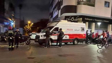 Night street scene with a red-and-white police van parked at a corner, officers and onlookers nearby at night.
