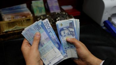 Two hands hold and count a fan of blue 200 denomination banknotes on a dark desk with a cash drawer and office printer in the background.