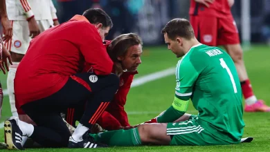 Soccer goalkeeper in green sits on the grass as medical staff and teammates check on him during a match nearby.