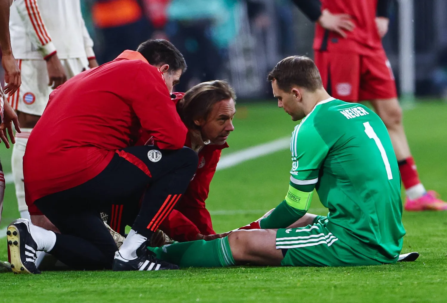 Soccer goalkeeper in green sits on the grass as medical staff and teammates check on him during a match nearby.
