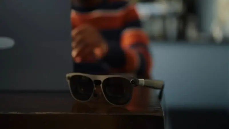 Sunglasses resting on a wooden desk with a blurred person using a laptop in the background.