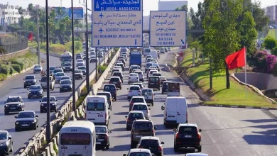 Busy Moroccan highway with many cars and two large overhead blue signs listing El Jadida and Marrakech in both Arabic and French.