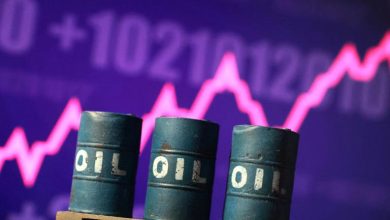 Three blue oil barrels labeled OIL in front of a purple stock market chart with a bright pink fluctuating line in the background.