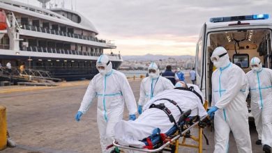 Medical team in full protective suits wheeling a patient on a stretcher toward an ambulance at a port, with a large cruise ship in the background.](https://)