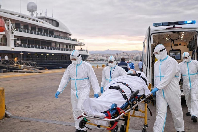 Medical team in full protective suits wheeling a patient on a stretcher toward an ambulance at a port, with a large cruise ship in the background.](https://)