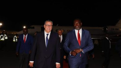 Two men in dark suits walk on a red carpet at night, with a private jet in the background.