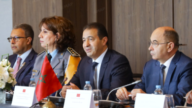 Four officials seated at a formal conference table with a Moroccan flag, microphones, and name placards; the man in the center is speaking.
