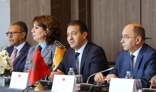 Four officials seated at a formal conference table with a Moroccan flag, microphones, and name placards; the man in the center is speaking.