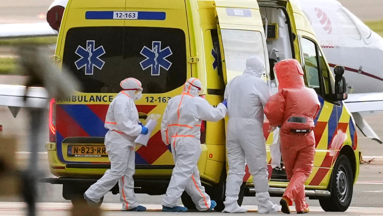 Medical responders in white hazmat suits help a person into a bright yellow ambulance on an airstrip, with a small airplane in the background.