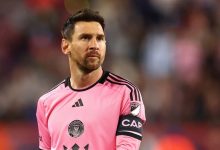 Male soccer goalkeeper in a pink kit with black stripes and captain's armband, standing on the field with a blurred crowd behind him.
