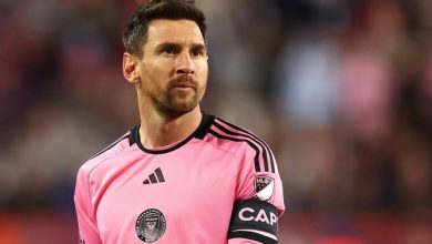 Male soccer goalkeeper in a pink kit with black stripes and captain's armband, standing on the field with a blurred crowd behind him.