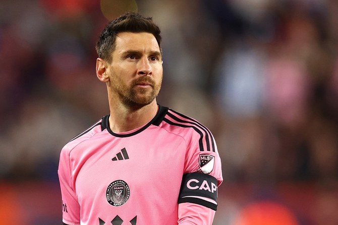 Male soccer goalkeeper in a pink kit with black stripes and captain's armband, standing on the field with a blurred crowd behind him.