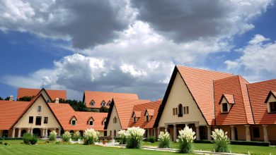Row of beige buildings with red-tiled, steeply pitched roofs and triangular gables set in a manicured lawn under a cloudy sky.
