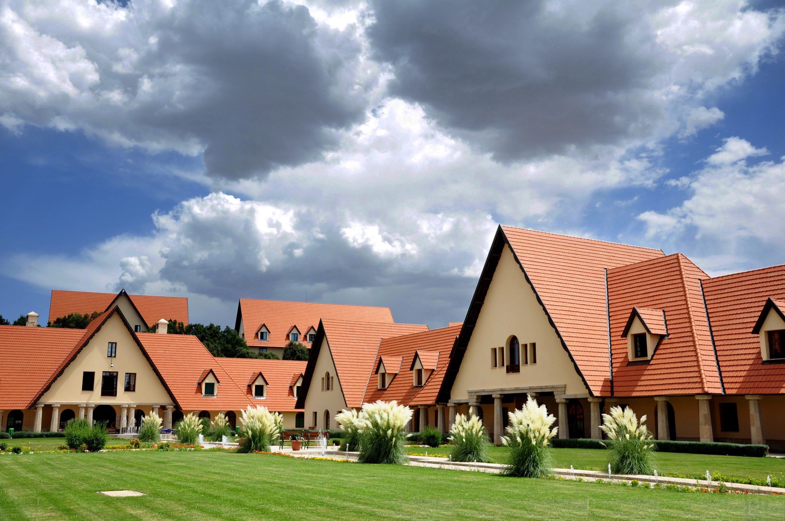 Al_Akhawayn_Campus | MCG24 | موقع إلكتروني مغربي شامل متجدد على مدار الساعة Row of beige buildings with red-tiled, steeply pitched roofs and triangular gables set in a manicured lawn under a cloudy sky.