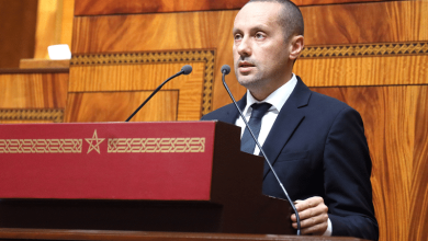 tahraoui | MCG24 | موقع إلكتروني مغربي شامل متجدد على مدار الساعة Man in a dark suit delivering a formal speech at a wooden podium with two microphones in a large wood-paneled chamber; calm, focused expression while addressing an audience.