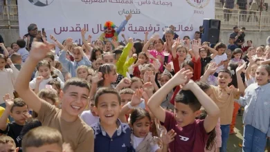 A large group of smiling children raise their hands at an outdoor event, with a banner and logos in Arabic behind them.