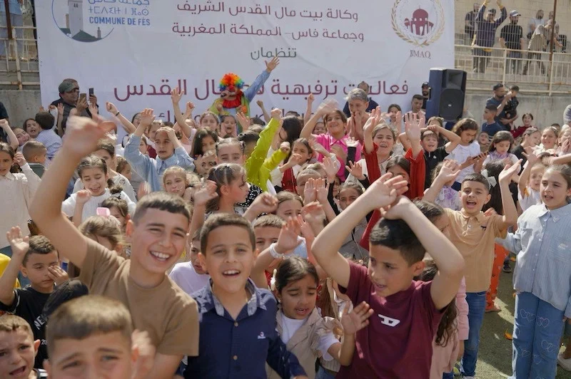 A large group of smiling children raise their hands at an outdoor event, with a banner and logos in Arabic behind them.