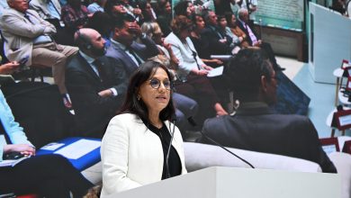 Female speaker in a white blazer at a lectern, delivering a speech with a large audience backdrop behind her.