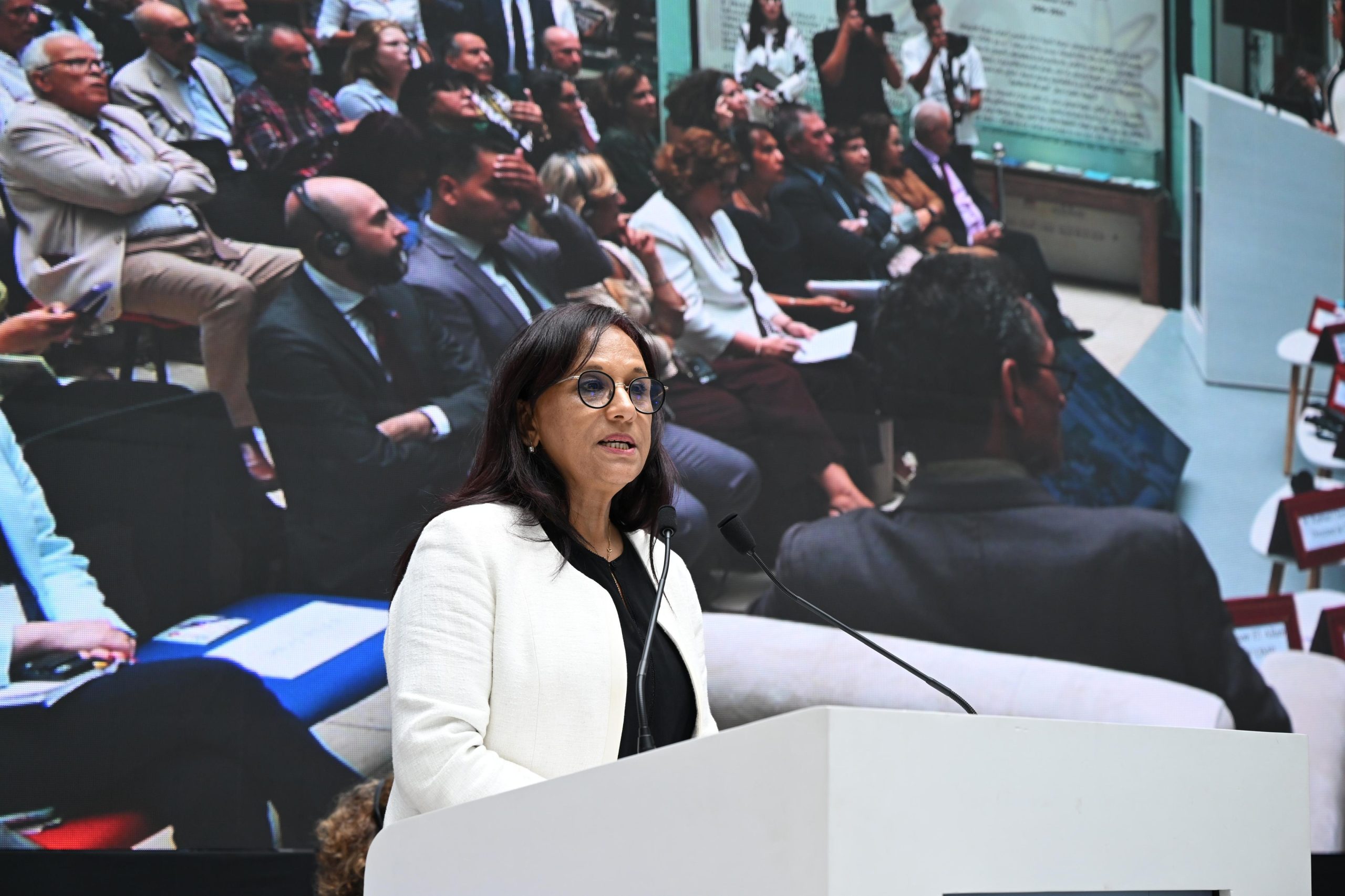 Female speaker in a white blazer at a lectern, delivering a speech with a large audience backdrop behind her.