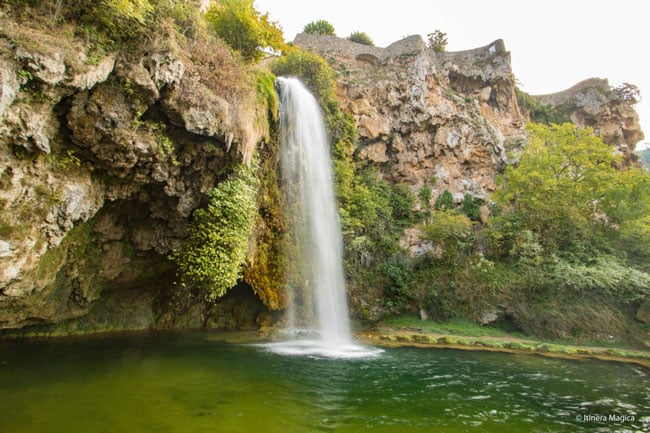 Tall waterfall flowing from a rocky cliff into a green pool, surrounded by mossy rocks and trees in a lush gorge.