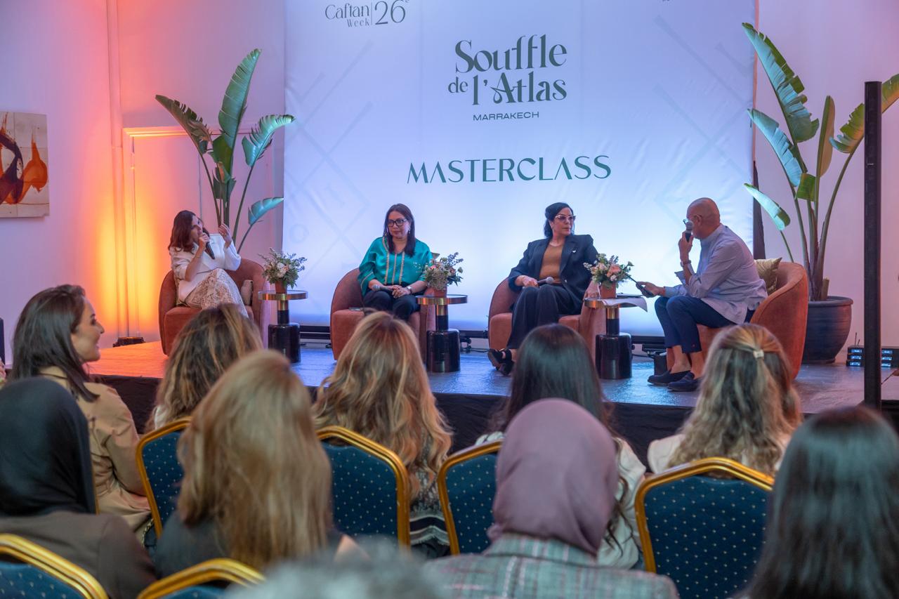 Panel discussion on a stage with four speakers seated in orange chairs, audience listening attentively.
