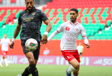 Two soccer players chase the ball on a green pitch; one in a dark jersey (Tanger Med), the other in white and red, near empty red‑green seats in the stadium.