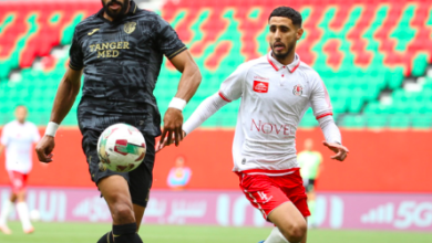 Two soccer players chase the ball on a green pitch; one in a dark jersey (Tanger Med), the other in white and red, near empty red‑green seats in the stadium.