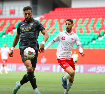 Two soccer players chase the ball on a green pitch; one in a dark jersey (Tanger Med), the other in white and red, near empty red‑green seats in the stadium.