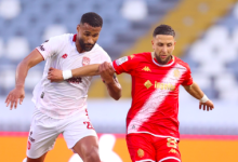 Two soccer players sprint for the ball during a match: a white-jersey opponent on the left and a red-jersey player on the right, with blurred stands in the background.