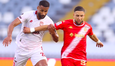Two soccer players sprint for the ball during a match: a white-jersey opponent on the left and a red-jersey player on the right, with blurred stands in the background.