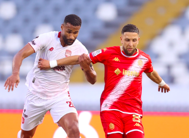 Two soccer players sprint for the ball during a match: a white-jersey opponent on the left and a red-jersey player on the right, with blurred stands in the background.