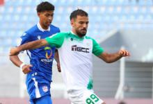 Two soccer players clash for the ball mid‑play: one in a white/green kit (number 99) and another in blue, with stadium seating in the background.