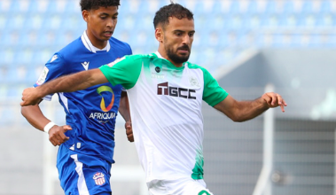 Two soccer players clash for the ball mid‑play: one in a white/green kit (number 99) and another in blue, with stadium seating in the background.