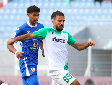 Two soccer players clash for the ball mid‑play: one in a white/green kit (number 99) and another in blue, with stadium seating in the background.