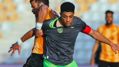 Soccer player in a gray jersey and neon green shorts (number 8) controls the ball as a defender in orange chases in a stadium.
