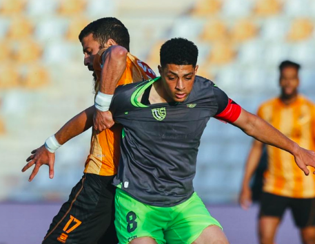 Soccer player in a gray jersey and neon green shorts (number 8) controls the ball as a defender in orange chases in a stadium.