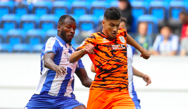 Two soccer players contest the ball during a match; one in blue-and-white stripes and the other in orange tiger-stripe kit, with blue seats in the stands behind them.