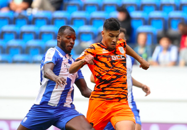 Two soccer players contest the ball during a match; one in blue-and-white stripes and the other in orange tiger-stripe kit, with blue seats in the stands behind them.
