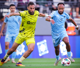 Two soccer players sprint for the ball in a match: one in a yellow kit and one in light blue.