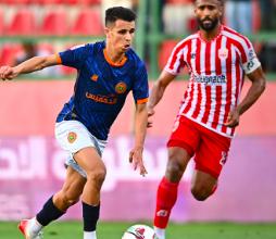 Two football players chase the ball on a green pitch during a match; one in a blue jersey and white shorts, the other in a red-and-white striped kit.