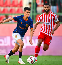 Two football players chase the ball on a green pitch during a match; one in a blue jersey and white shorts, the other in a red-and-white striped kit.