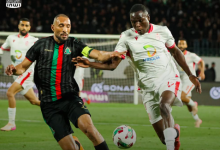 Two soccer players compete for the ball during a night match; one wears black with a green/red stripe, the other in white.