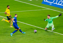 Soccer scene: a blue team player shoots toward goal as the goalkeeper in green dives to save, with a yellow-clad defender nearby on a green pitch and a ball rolling toward the net.