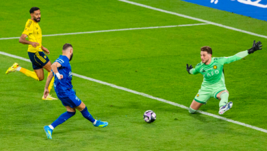 Soccer scene: a blue team player shoots toward goal as the goalkeeper in green dives to save, with a yellow-clad defender nearby on a green pitch and a ball rolling toward the net.