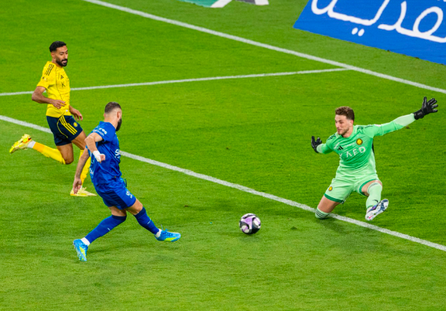 Soccer scene: a blue team player shoots toward goal as the goalkeeper in green dives to save, with a yellow-clad defender nearby on a green pitch and a ball rolling toward the net.