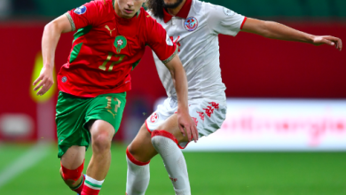 Two soccer players chase the ball during a match; one in red‑green kit, the other in white, on a green field.
